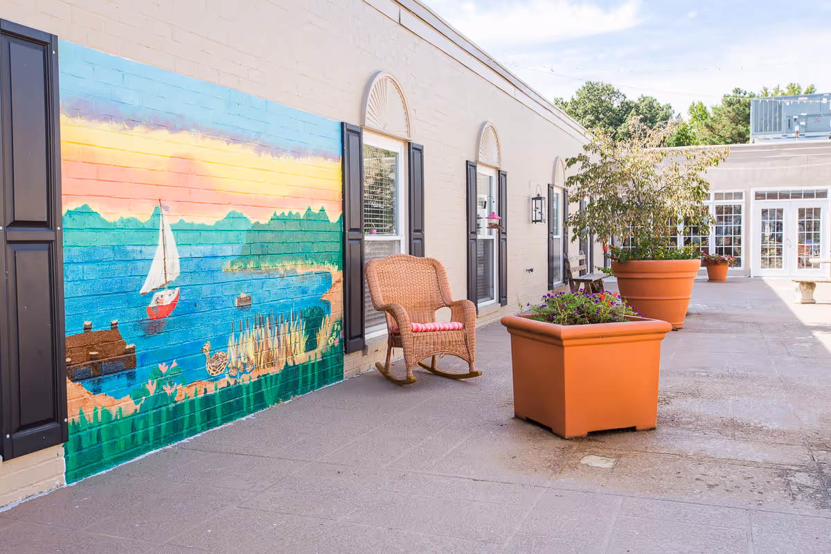 Outdoor patio area at Mallard Bay Nursing and Rehab with a colorful mural of a lake scene featuring a sailboat and ducks on a brick wall, a wicker rocking chair with a cushion, and large orange planters with flowers and small trees along the walkway.