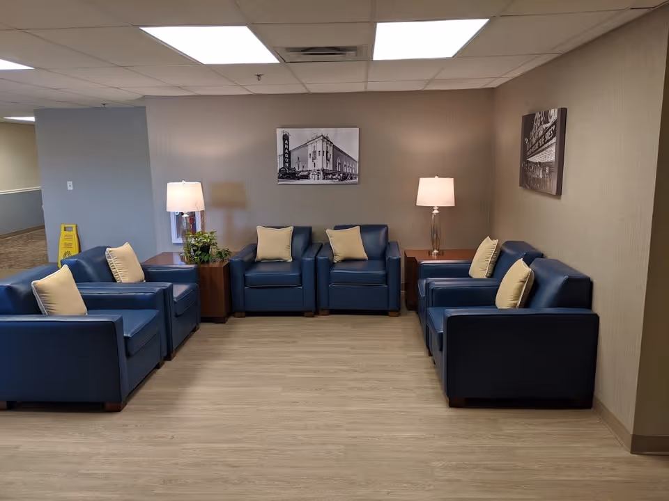 A seating area in a supportive living facility with six blue armchairs arranged in a U-shape, each with a light-colored pillow. There are two wooden side tables with lamps and a small green plant. The walls are beige with two black and white framed photographs hanging. The floor is light wood, and the ceiling has white tiles with fluorescent lighting.