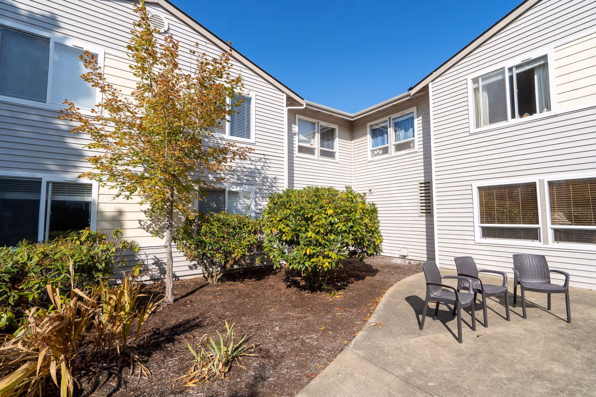 Outdoor courtyard area of a retirement facility with beige siding buildings, several windows, a small tree, green bushes, and three black plastic chairs arranged on a concrete patio under a clear blue sky.