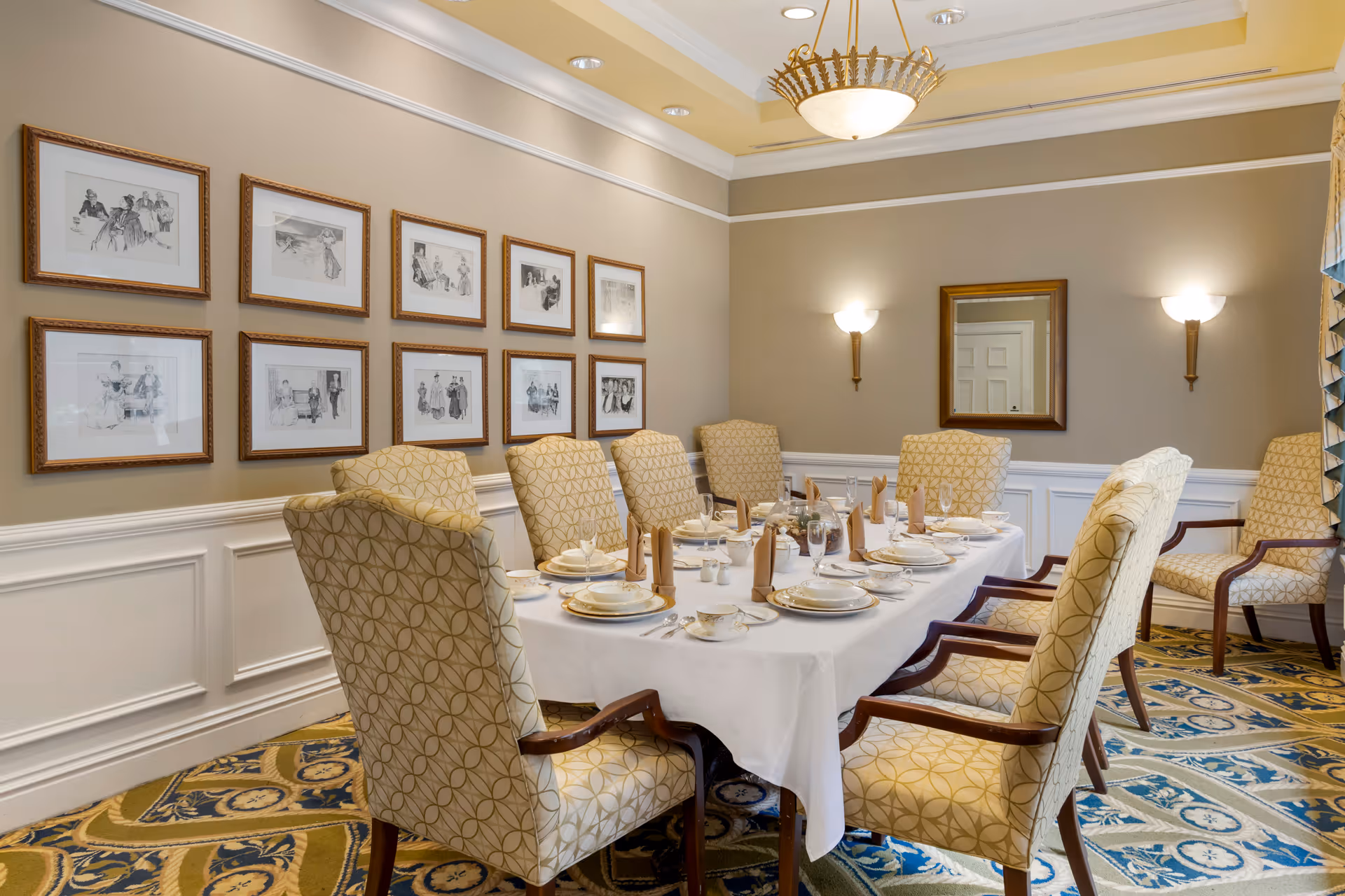 A formal dining room with a long table covered with a white tablecloth set for a meal. The table is surrounded by eight upholstered chairs with a geometric pattern. The walls are beige with white wainscoting and decorated with framed black and white sketches. Two wall sconces and a chandelier provide lighting, and a mirror hangs on the far wall.