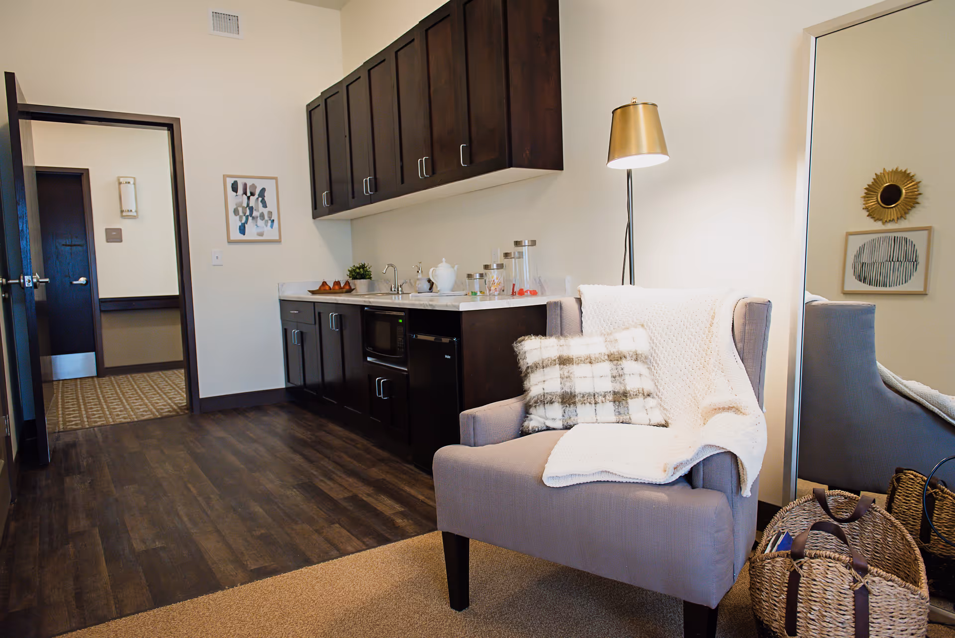 A cozy interior room featuring a gray armchair with a plaid pillow and a white knitted throw blanket. Next to the chair is a tall floor lamp with a gold lampshade. Behind the chair is a large mirror reflecting part of the room. To the left, there is a kitchenette area with dark wood cabinets, a small sink, a microwave, and a mini fridge. The floor is a combination of dark wood and carpet. The walls are light-colored with framed artwork and a doorway leading to another room.