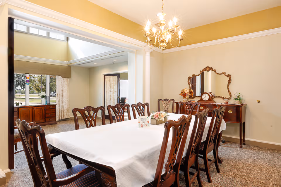 A well-lit dining room with a long rectangular table covered with a white tablecloth and surrounded by wooden chairs with intricate backs. A chandelier hangs above the table. Against the wall is a wooden sideboard with a decorative mirror, a clock, and floral arrangements. Large windows in the adjacent room let in natural light, and the walls are painted in soft beige tones.