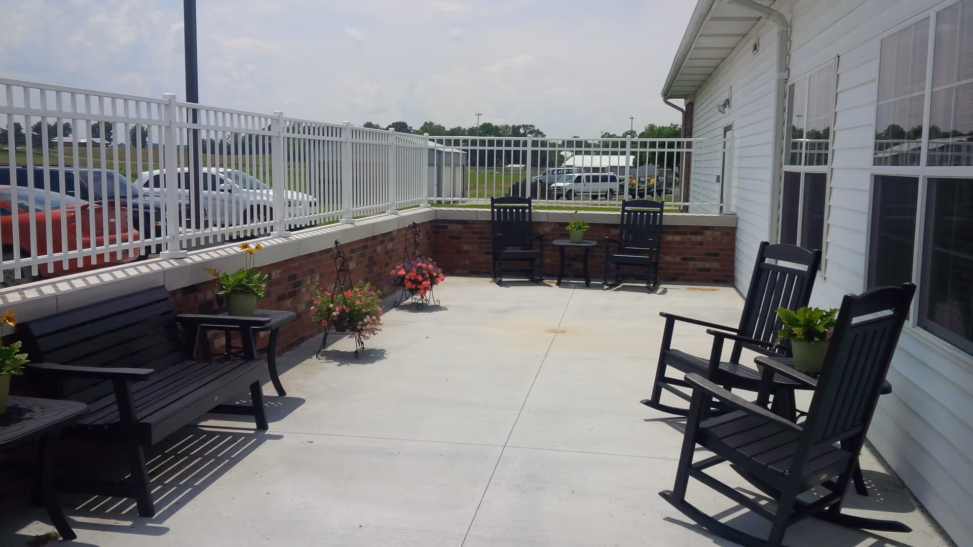 Outdoor patio area with black wooden benches and rocking chairs arranged along a white building wall and a brick half-wall with white metal fencing. Several potted plants and flowers are placed on small tables and stands around the seating area. Cars and green fields are visible beyond the fence under a partly cloudy sky.