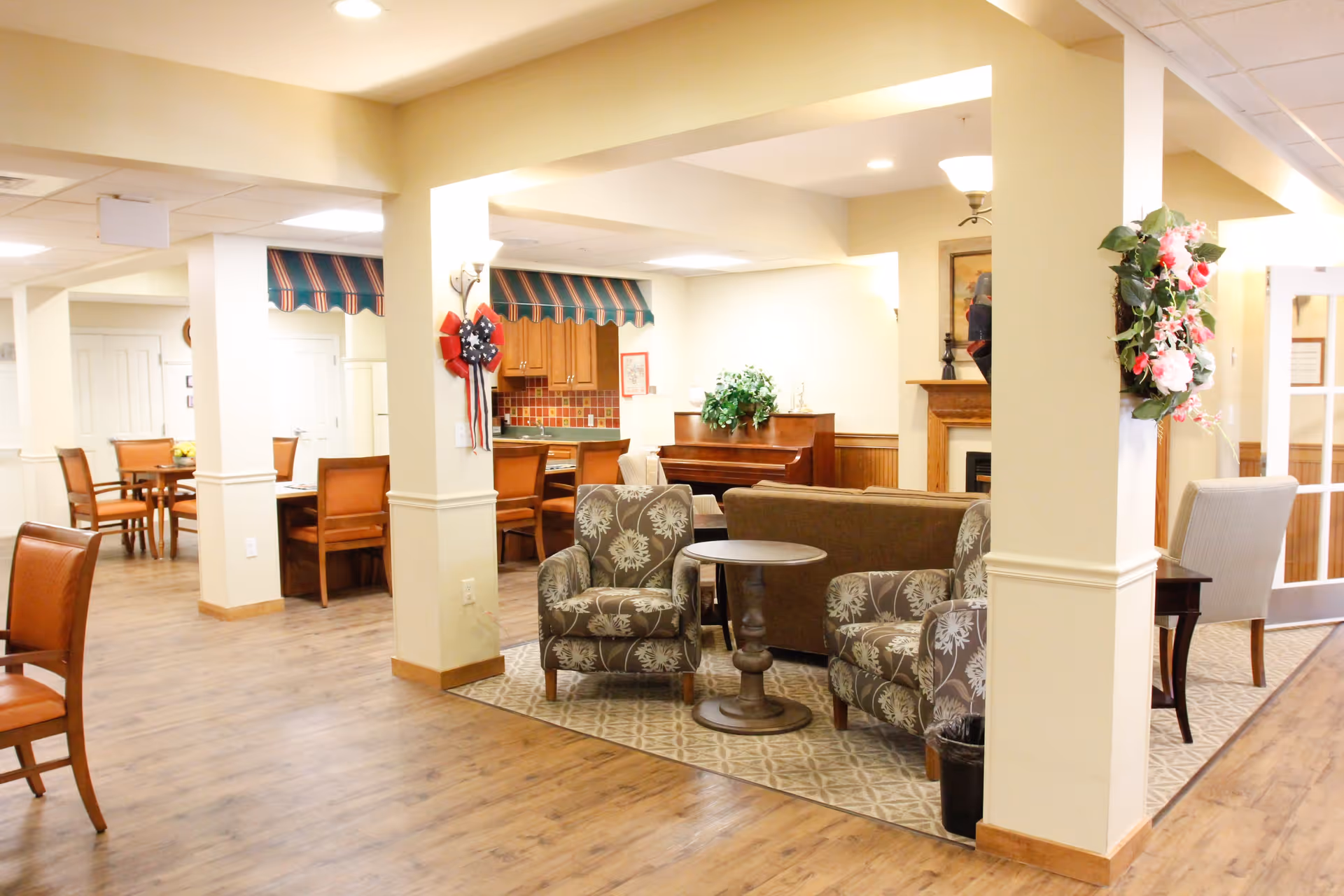 A bright and spacious common area in a senior living facility featuring a seating area with two patterned armchairs and a small round table on a patterned rug, a brown sofa, a piano against the wall, and a dining area with wooden chairs and tables. The room has light-colored walls, wood flooring, decorative floral wreaths on columns, and a fireplace in the background.