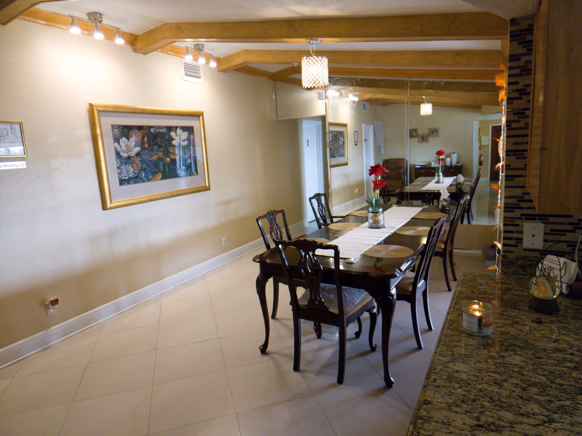 A dining area with a long wooden table and six chairs. The table has a white runner and a vase with red flowers. The room features beige walls, a large mirror on one side, a framed floral painting, and wooden ceiling beams with track lighting. A granite countertop with a lit candle is visible in the foreground.