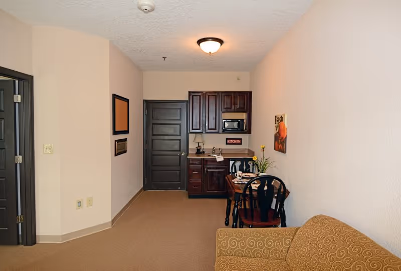 Interior view of a small living space in Webb House Retirement Center featuring a kitchenette with dark wood cabinets, a microwave, and a small dining table with two chairs. A patterned armchair is partially visible in the foreground, and there are two dark doors on the left side of the room. The walls are light beige, and the floor is carpeted.