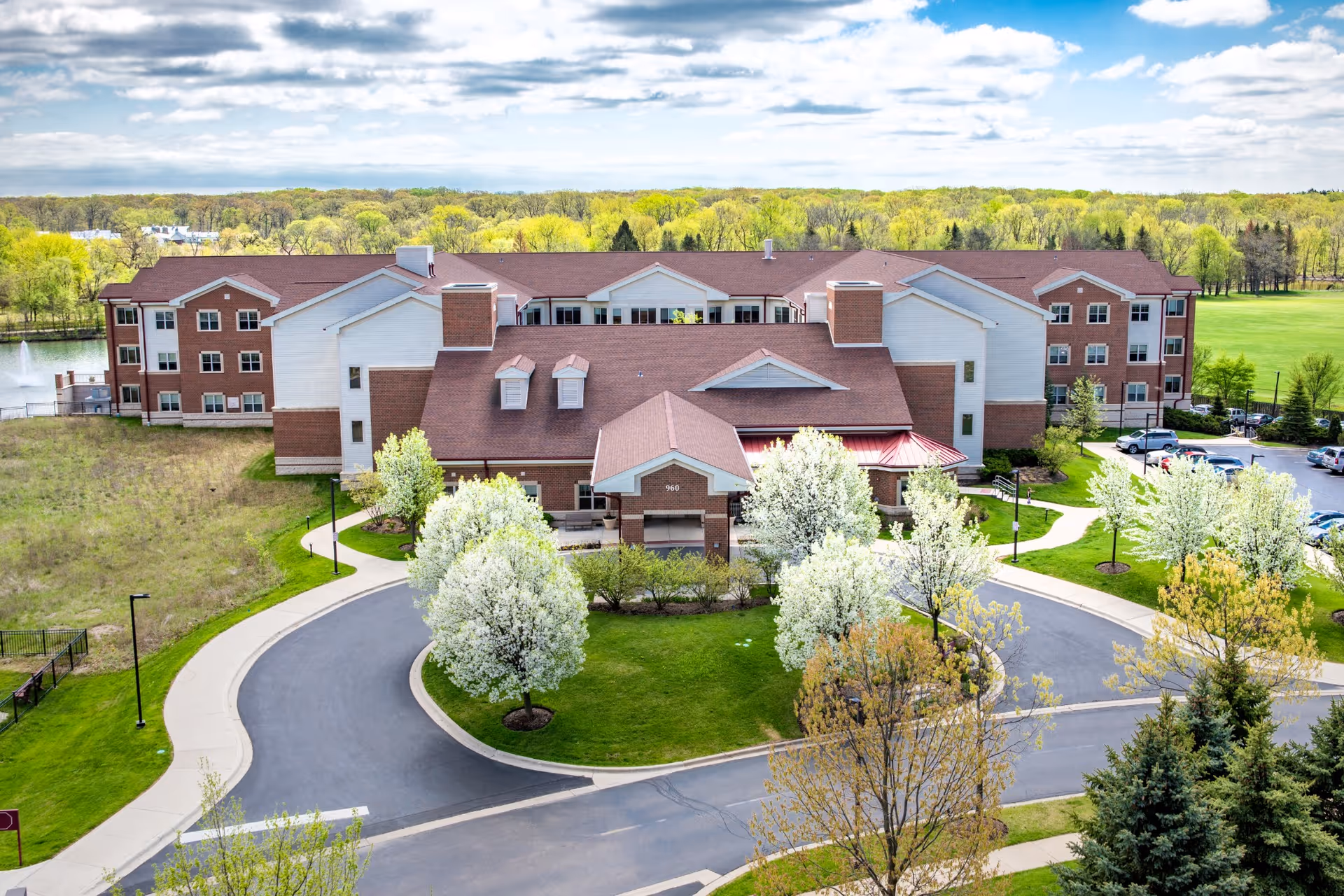 A large senior living facility building with a red roof and white and brick exterior surrounded by green lawns and blooming trees. A curved driveway leads to the main entrance, and there is a parking lot visible on the right side. The background features a wooded area under a partly cloudy sky.
