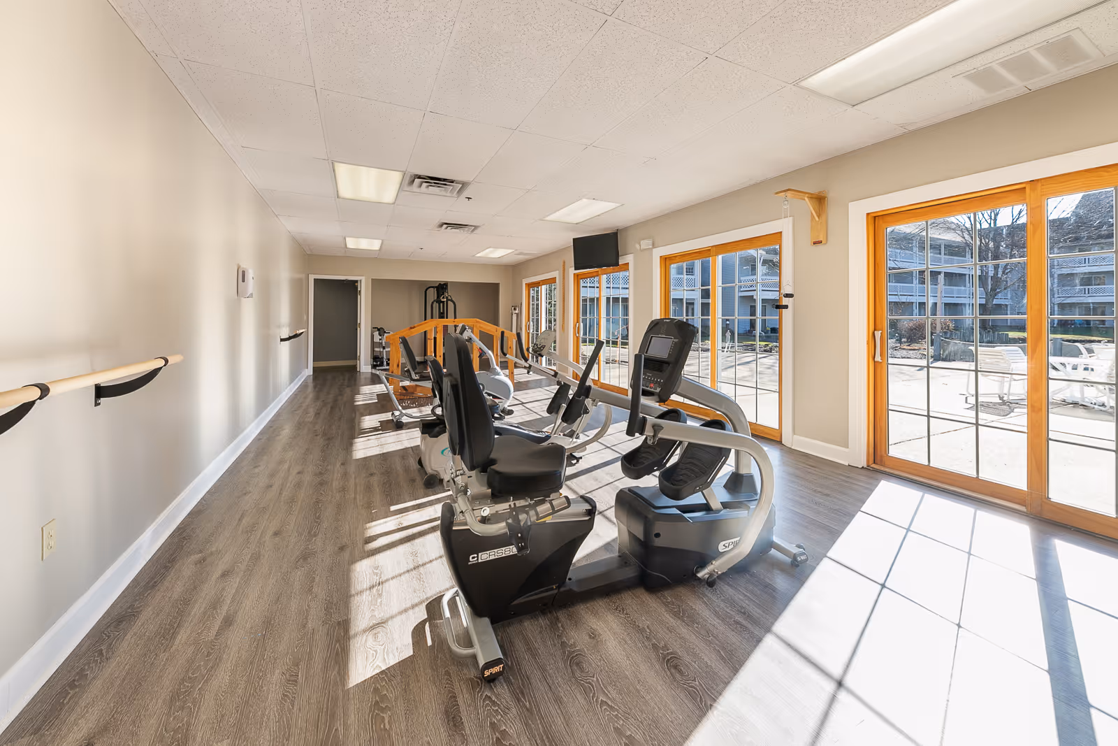 Bright exercise room with large windows letting in natural light, featuring stationary recumbent bikes and other fitness equipment. The room has wood flooring, beige walls with handrails, and a TV mounted on the wall. Outside the windows, there is a patio area with chairs and tables.
