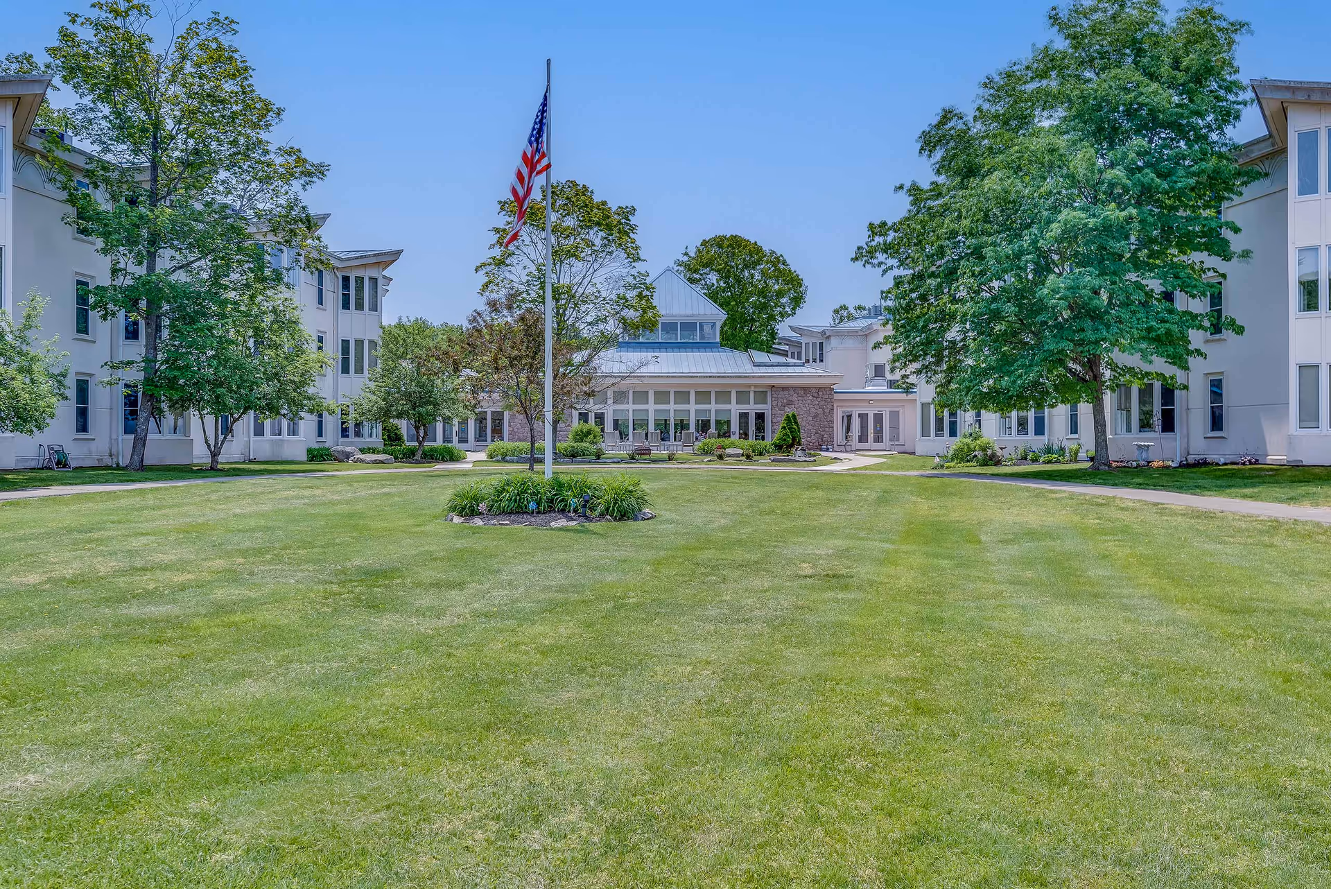 A large, well-maintained grassy courtyard with a flagpole flying the American flag in the center. The courtyard is surrounded by a white multi-story building with many windows and trees providing shade. The sky is clear and blue.