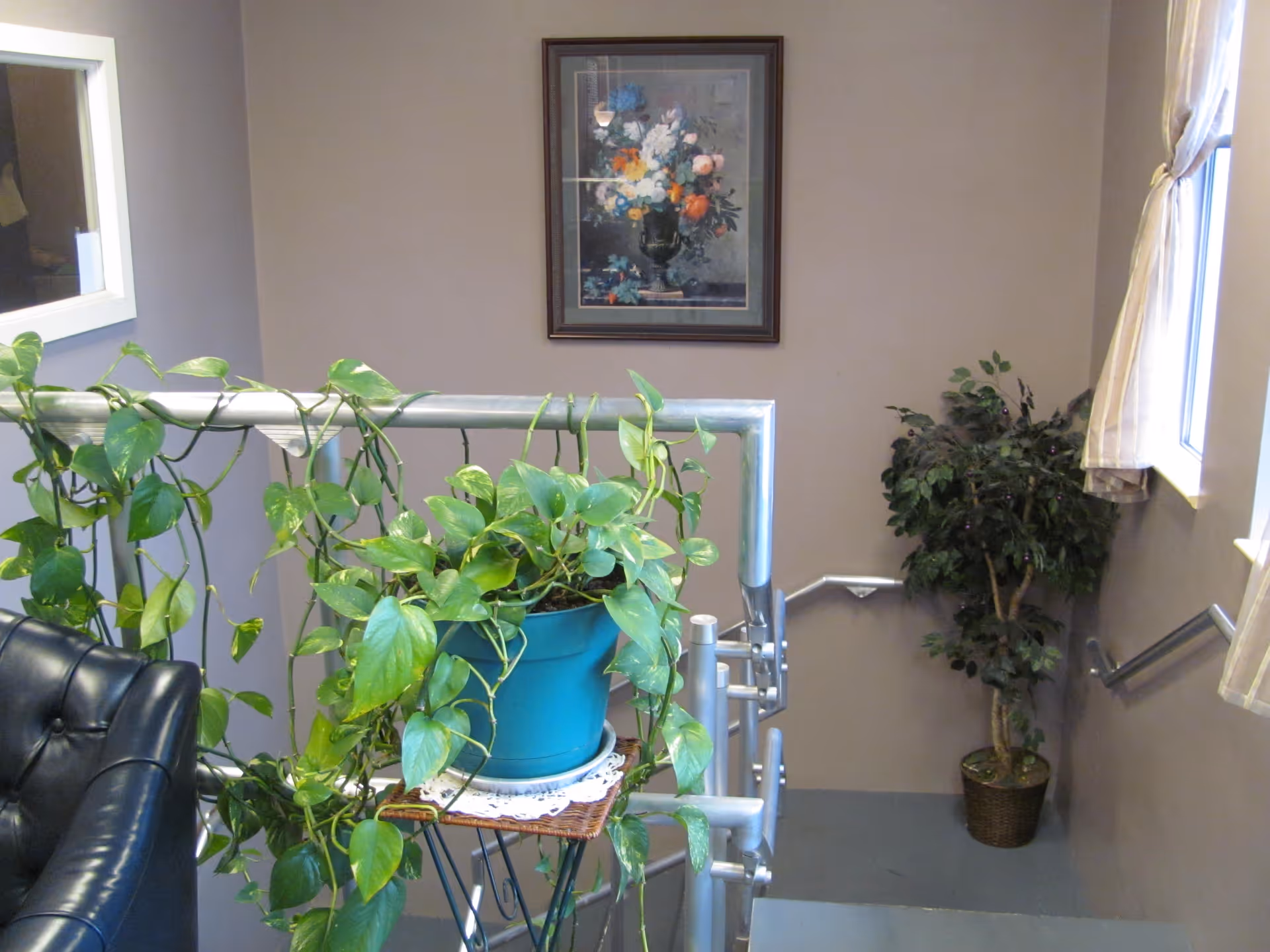 Indoor stairwell area with a metal handrail decorated with a green potted plant in a blue pot on a small stand. A black leather chair is partially visible on the left. There is a framed floral painting on the beige wall above the stairs, and a window with beige curtains on the right side. Another potted plant is placed on the floor near the window.