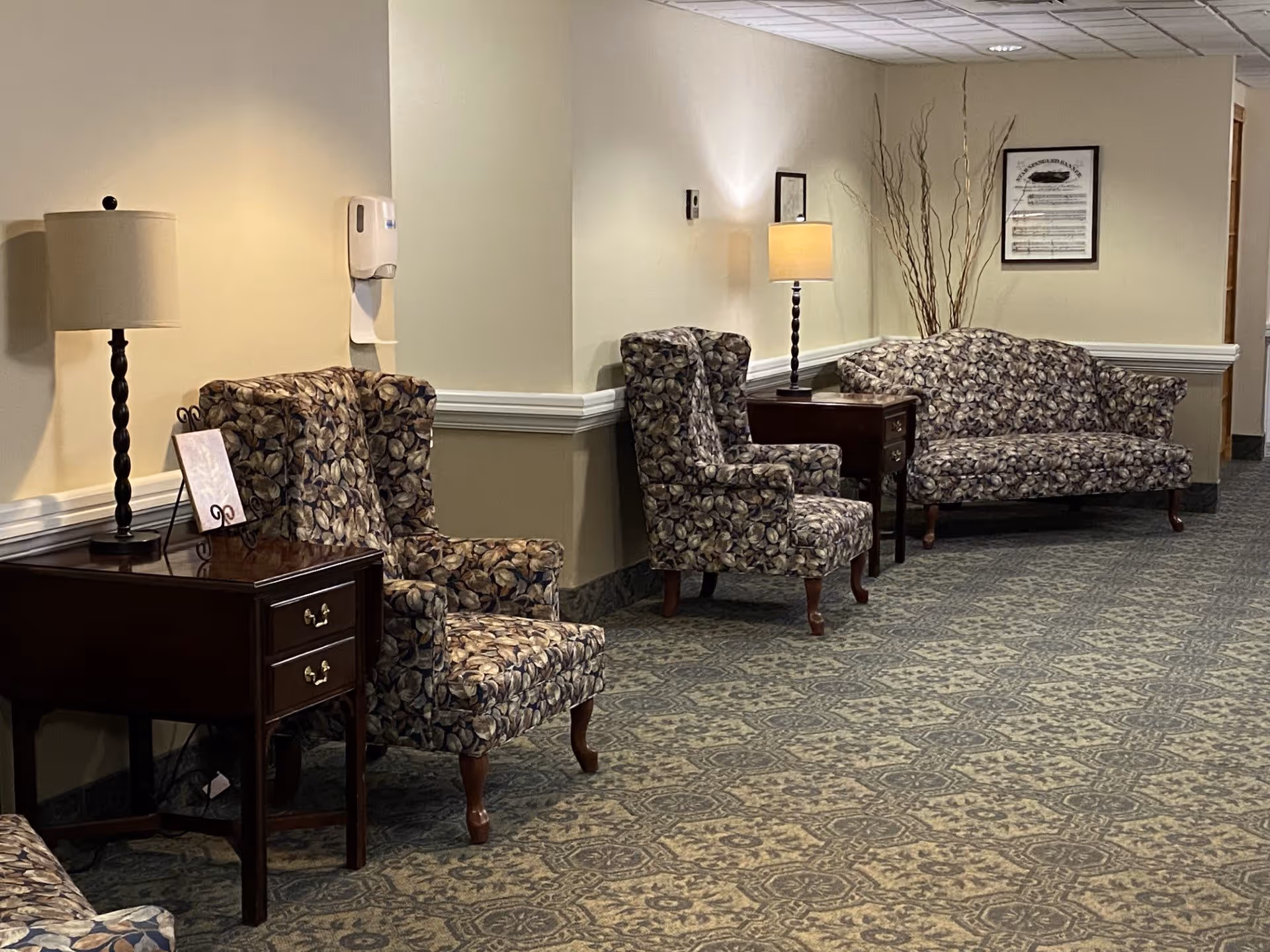 A hallway seating area in a senior living facility with patterned armchairs and a matching loveseat. There are two wooden side tables with lamps on them, a framed picture on the wall, and decorative branches in a vase. The carpet has a detailed pattern and the walls are painted in a light beige color with white trim.