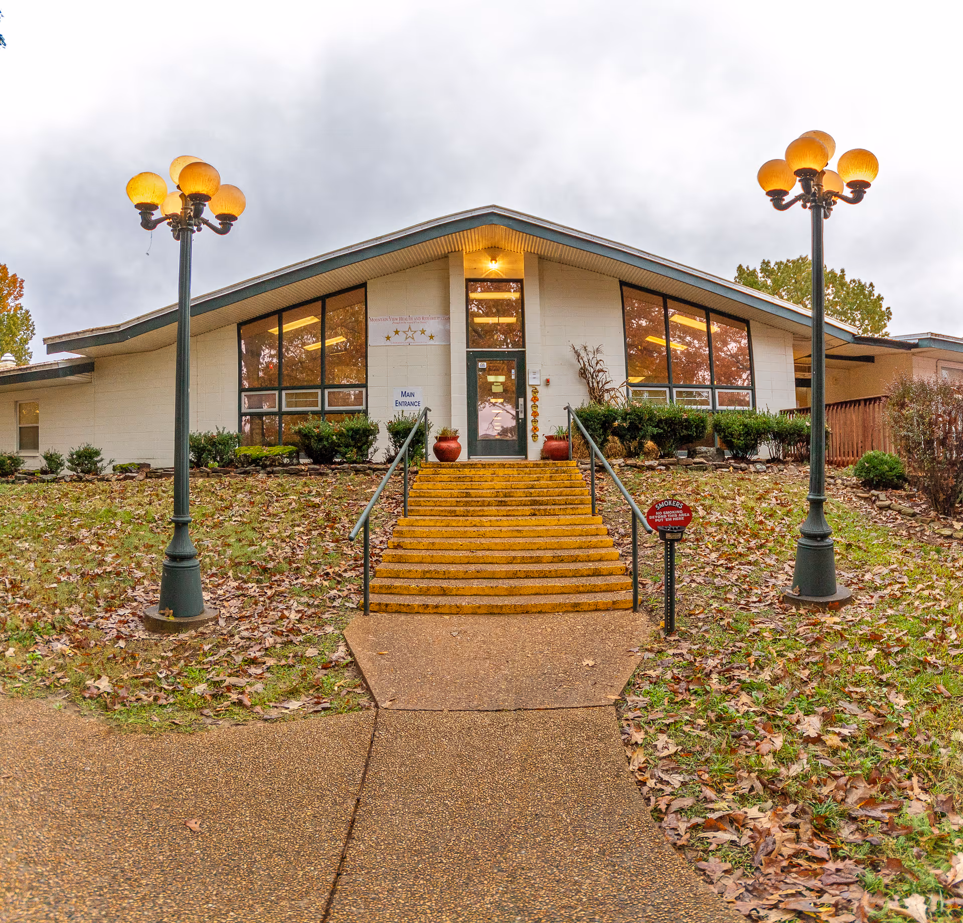 Front entrance of a single-story nursing facility with steps leading up to double doors, flanked by lamp posts and large windows.