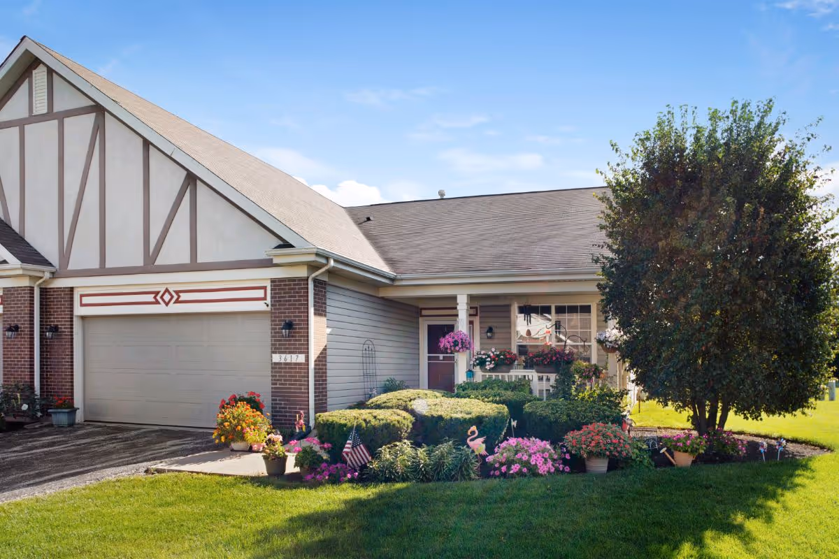 Front exterior of a ranch-style residential building with an attached garage, a flower-adorned porch, and a manicured lawn and garden.