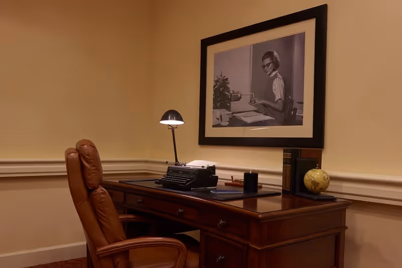 A traditional office corner with a wooden desk and a brown leather chair. On the desk, there is a vintage typewriter, a small desk lamp, some books, a globe, and a few office supplies. A framed black and white photograph of a woman working at a typewriter hangs on the beige wall above the desk.