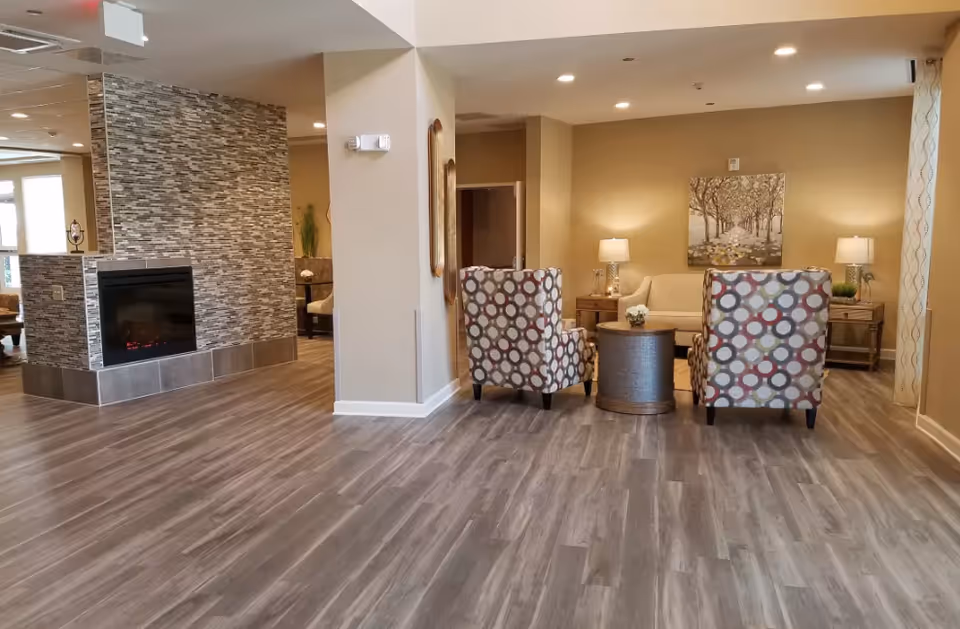 A cozy living room area with two patterned armchairs facing a beige sofa. A round wooden coffee table is placed between the chairs and sofa. The room features warm beige walls, two side tables with lamps, a painting of a tree-lined path on the wall, and a modern fireplace with a tiled surround. The floor is covered with wood-like vinyl planks.