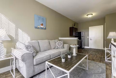 A bright living room area with a light gray sofa adorned with patterned pillows, a wooden coffee table with white metal legs holding small potted plants, a side table with a lamp, and a painting of a dog on the wall. The room has wood flooring and leads to a small kitchen area with a refrigerator and microwave. The entrance door is visible in the background.