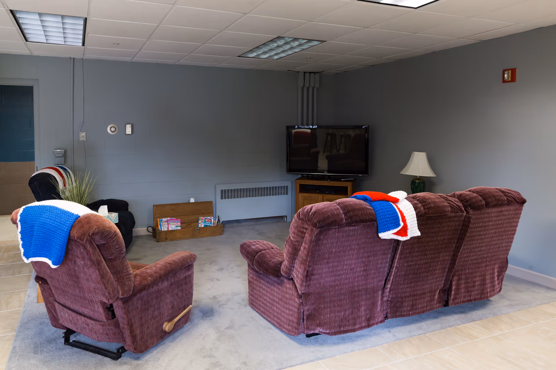 A cozy senior community living room with a maroon recliner and matching three-seater sofa, both draped with red, white, and blue knitted blankets. A flat-screen TV is mounted on a wooden stand against a gray wall. There is a lamp on a side table next to the sofa, a wooden box with magazines on the floor, and a small black armchair in the corner. The room has a tiled floor with a carpeted area under the seating.