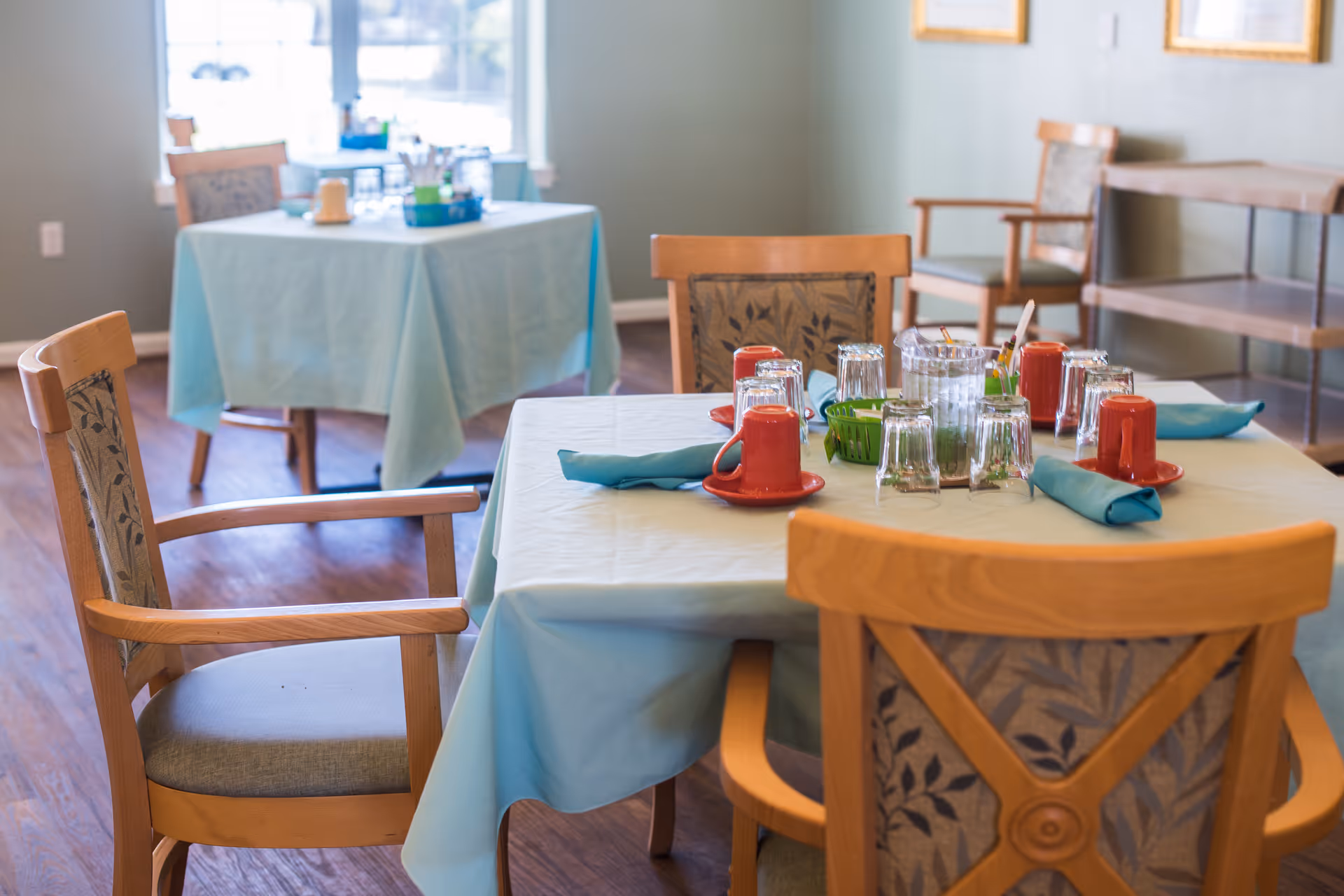A dining room in a senior living community with wooden chairs around tables covered with light blue tablecloths. The tables are set with red coffee cups, glasses, napkins, and a pitcher of water. The room has wooden flooring and light-colored walls with framed artwork.