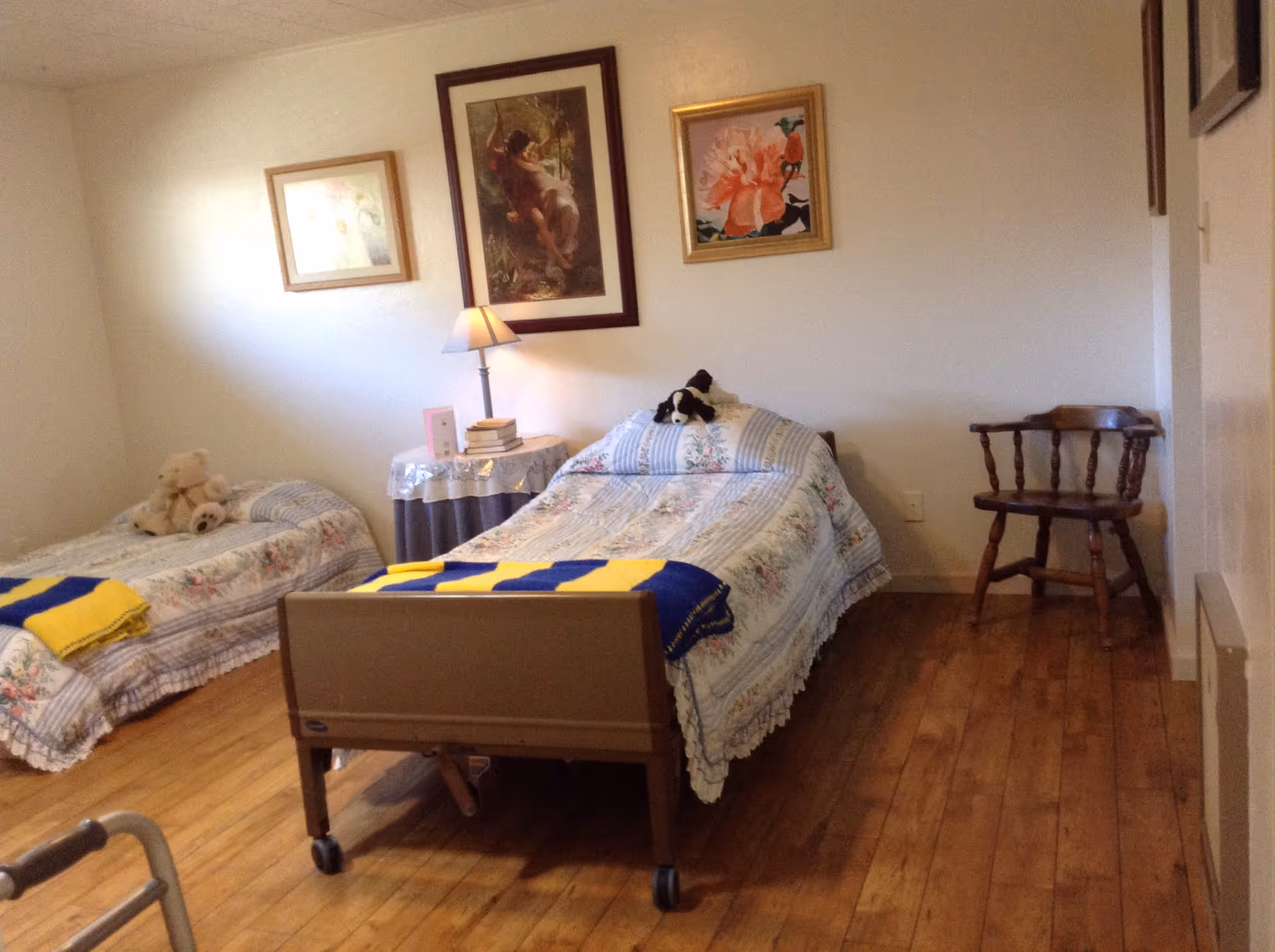 A bedroom with two single beds covered in floral bedspreads, each with a stuffed animal on top. A small round table with a lamp and books is placed between the beds. Three framed pictures hang on the wall above the beds. There is a wooden chair in the corner and a walker partially visible in the foreground. The floor is wooden.