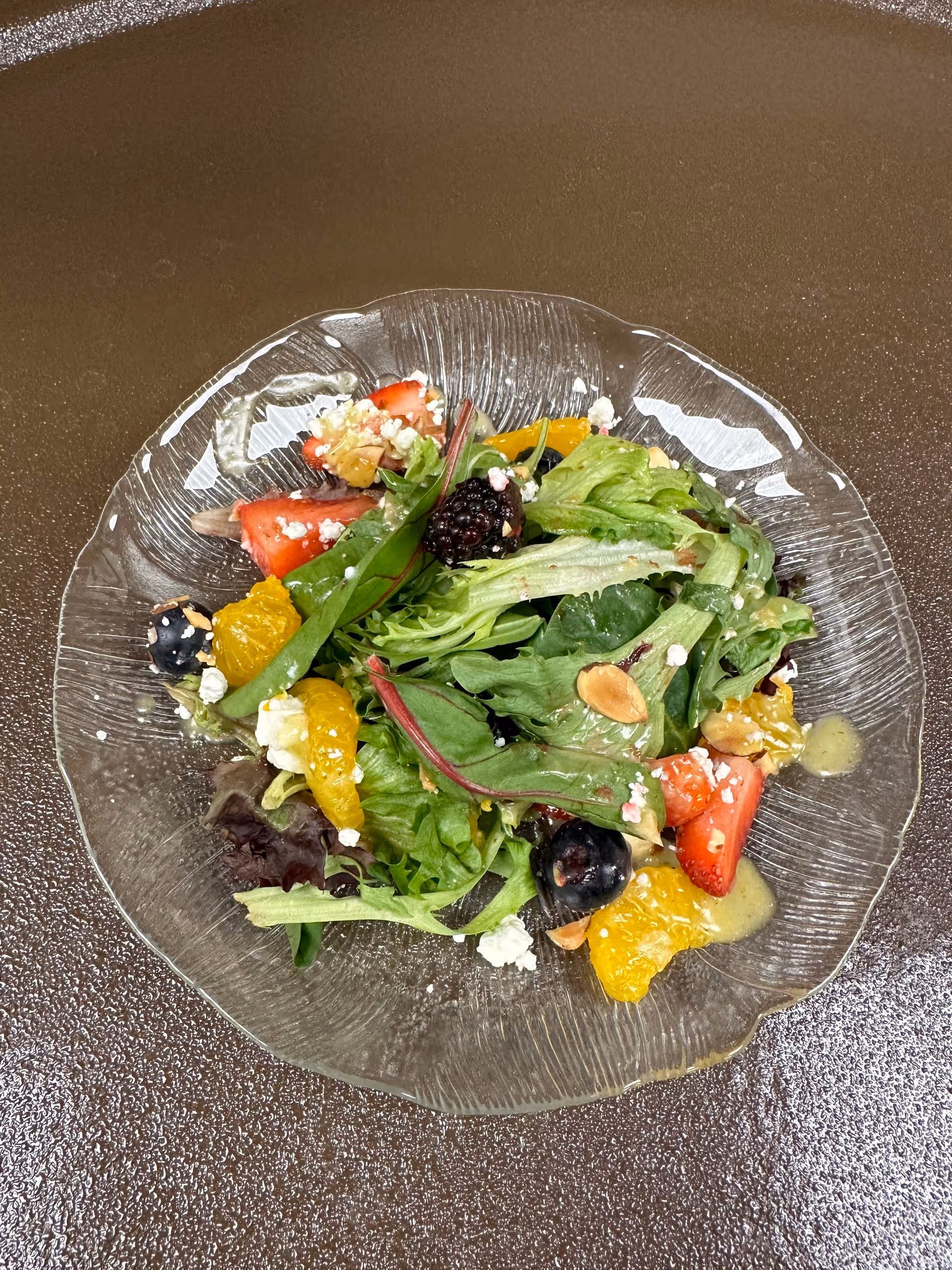 A clear glass plate with a fresh mixed green salad topped with sliced strawberries, blackberries, blueberries, mandarin orange segments, crumbled cheese, and sliced almonds, placed on a brown textured surface.