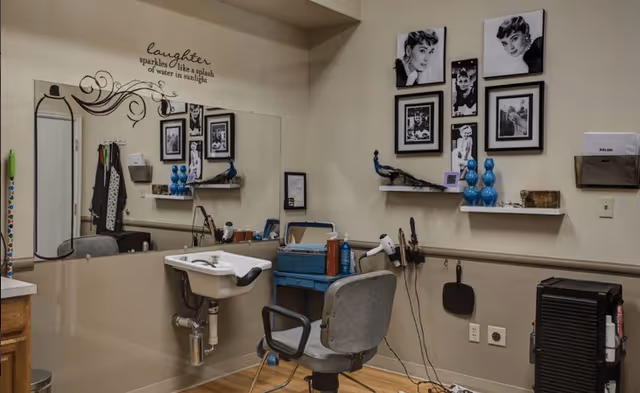 Interior view of a small salon or grooming area with a salon chair, a white sink, a large mirror, and shelves on the wall holding decorative items and framed black and white photos. The wall has a quote that reads 'laughter sparkles like a splash of water in sunlight.' Various hair styling tools and products are visible on a blue cart and hanging on the wall.