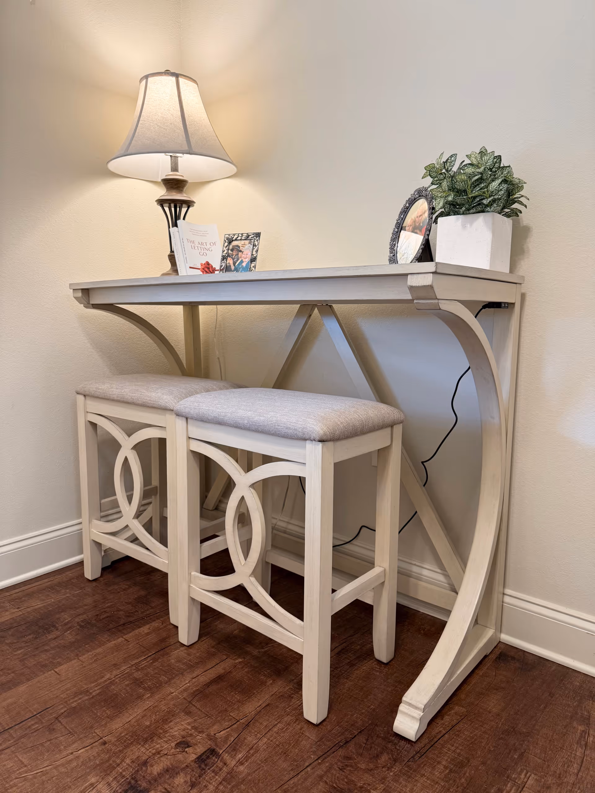 A small white wooden console table with two matching cushioned stools underneath, placed against a beige wall on a wooden floor. On the table, there is a lit table lamp, a small potted plant, a framed photo, and a book titled 'The Art of Letting Go'.