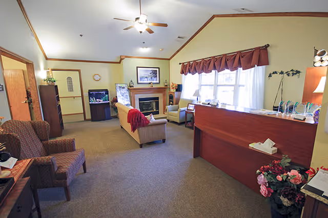 A cozy living room area in a senior living facility with beige walls and carpeted floor. The room features a ceiling fan with lights, a fireplace with a framed picture above it, a fish tank, and comfortable seating including armchairs and a sofa with a red throw blanket. A wooden reception desk is positioned near a window with brown valances, and there are decorative flowers and various items on the desk and around the room.