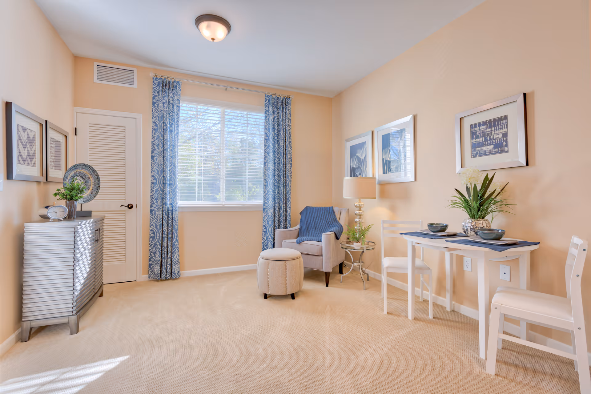 A bright and cozy room with beige walls and carpet, featuring a window with blue patterned curtains. The room includes a small white dining table set with two chairs, a beige armchair with a matching ottoman, a side table with a lamp and plant, and a silver chest with decorative items. Three framed artworks hang on the walls.