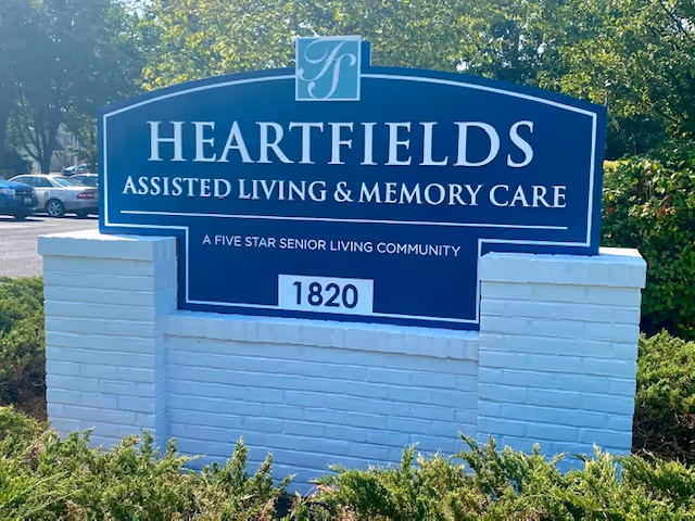 A blue and white sign for Heartfields Assisted Living & Memory Care, a five star senior living community, with the number 1820 displayed. The sign is mounted on a white brick base and surrounded by greenery, with trees and parked cars visible in the background.