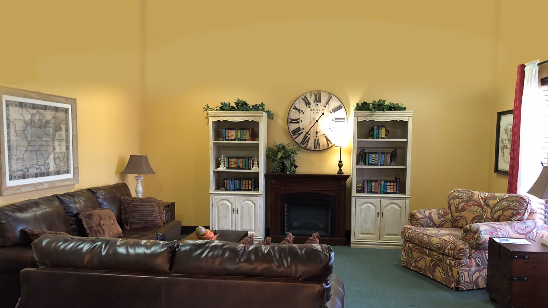 A cozy living room with yellow walls featuring a dark brown leather sofa set, a patterned armchair, two white bookshelves filled with books, a large round wall clock above a dark wooden electric fireplace, and a side table with a lamp.