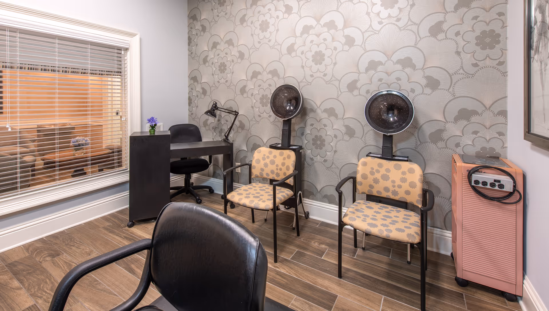 Interior room with two beige chairs with floral patterns placed under two black hair dryers, a black desk with a black swivel chair, a small pink cabinet with electrical outlets, and a window with white blinds showing a seating area with a couch and table with flowers behind it. The wall has a large floral patterned wallpaper and the floor is wood-tiled.