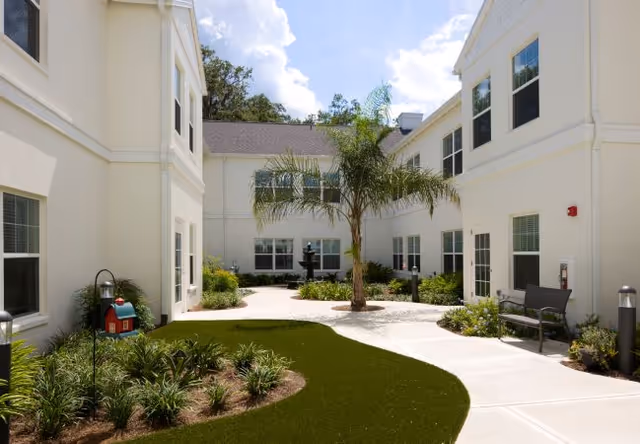 Sunlit courtyard with a central palm tree and fountain surrounded by two-story cream buildings, pathways, benches, and landscaping.