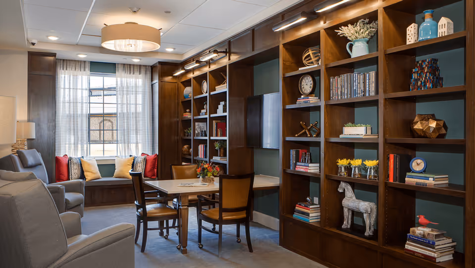 A cozy living room area with a large window featuring sheer curtains, a built-in window seat with cushions, two gray armchairs, a table with four chairs, and a large wooden bookshelf filled with books, decorative items, and plants. The room is softly lit with ceiling lights and a hanging lamp.