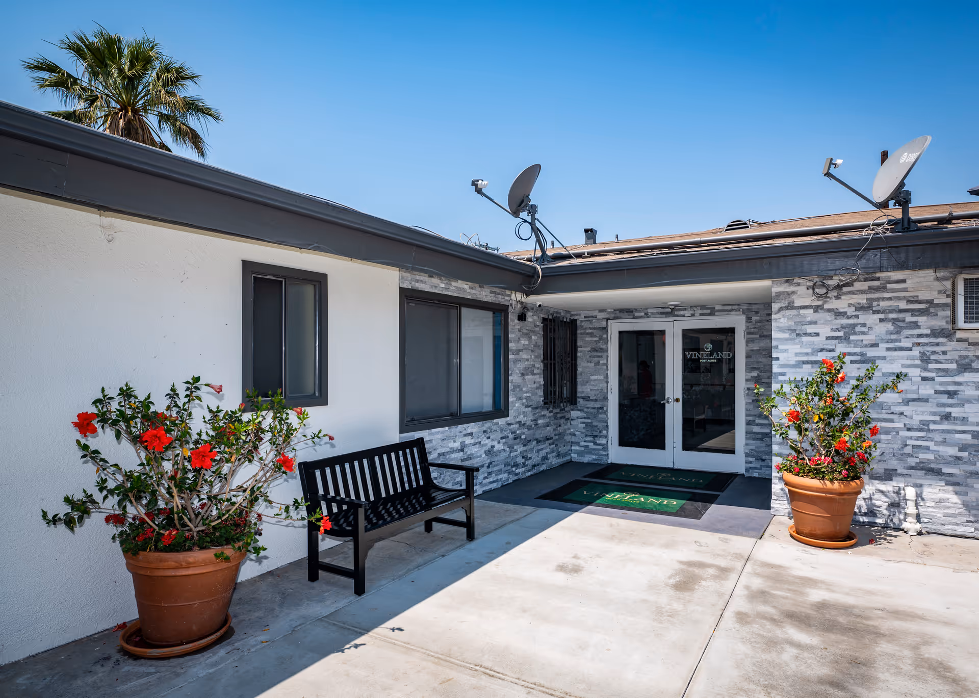 Entrance courtyard of Vineland Post Acute with double glass doors, a bench, potted flowering plants, and satellite dishes on the roof.