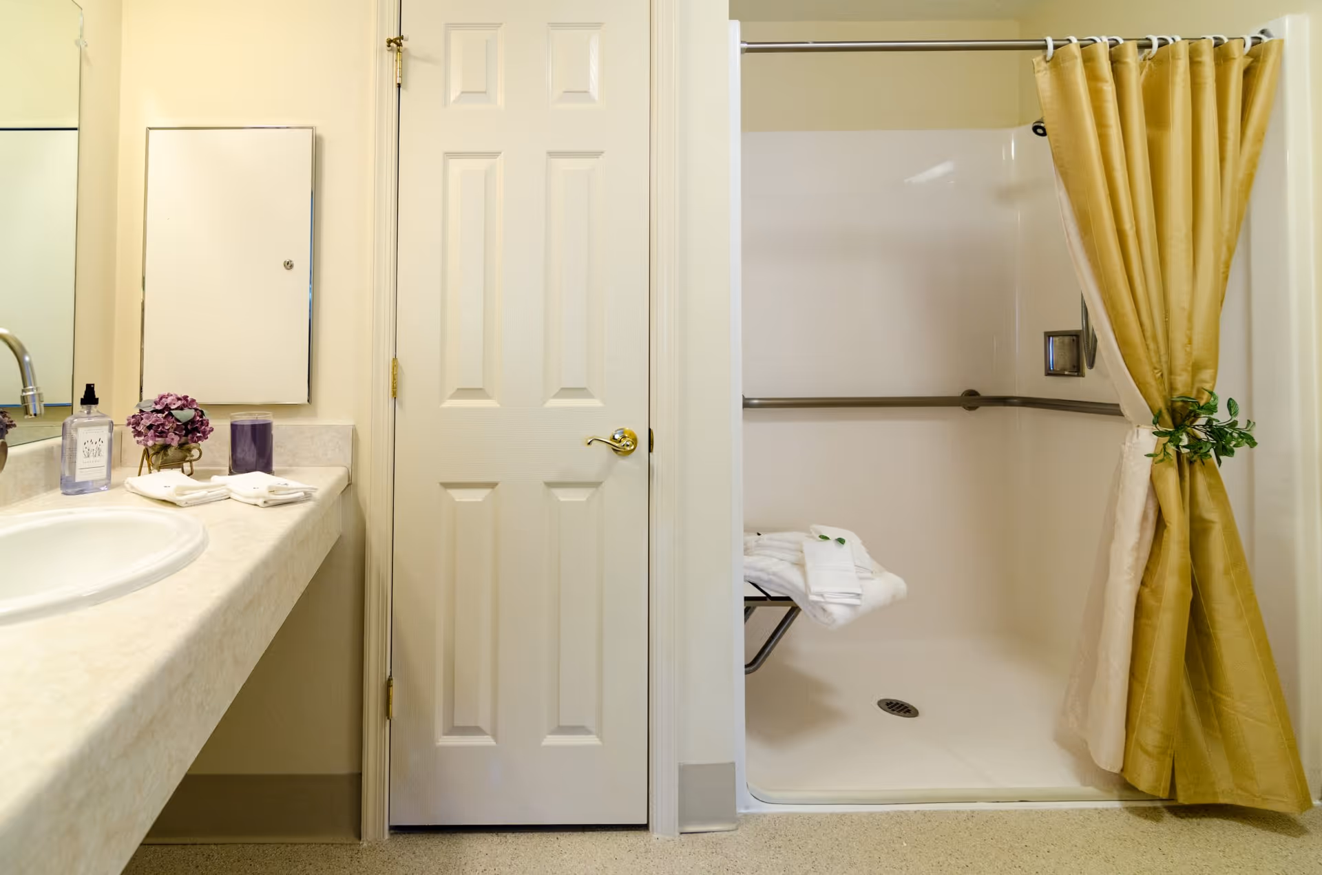 A clean bathroom with a sink and mirror on the left, a closed door in the center, and a walk-in shower with a tied-back curtain, grab bars, and a folded towel on a bench on the right.