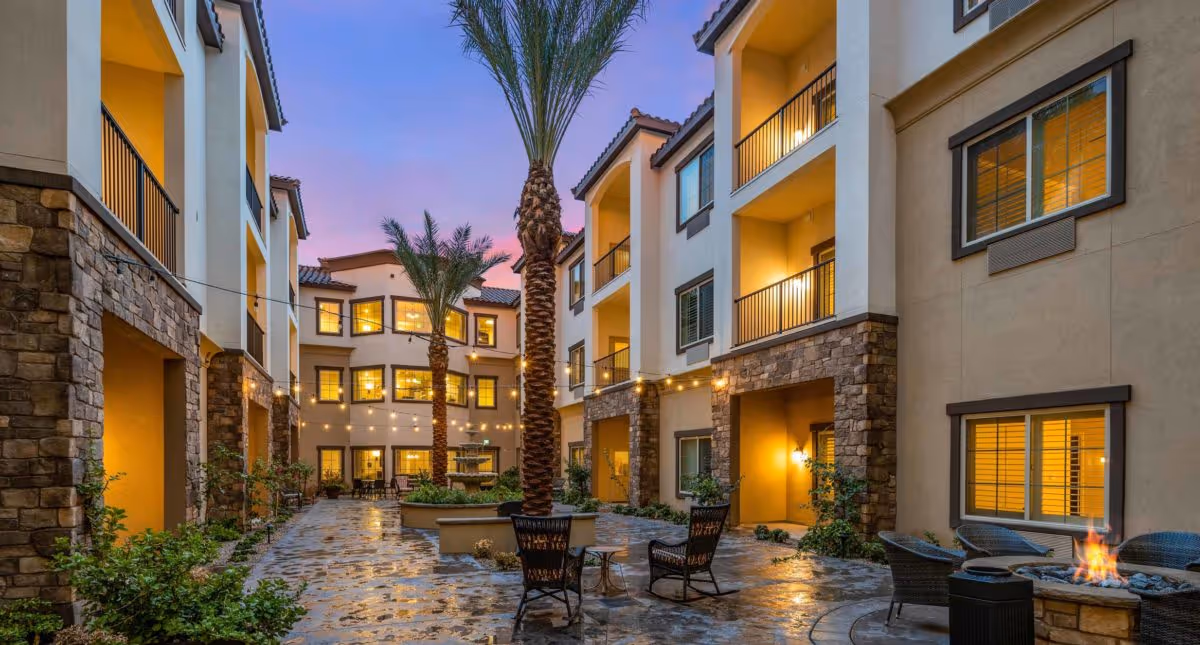 Outdoor courtyard area of The Park at Modesto Independent Living Community during dusk, featuring stone pathways, palm trees, string lights, a central fountain, and seating areas including chairs and a fire pit, surrounded by multi-story residential buildings with balconies and lit windows.