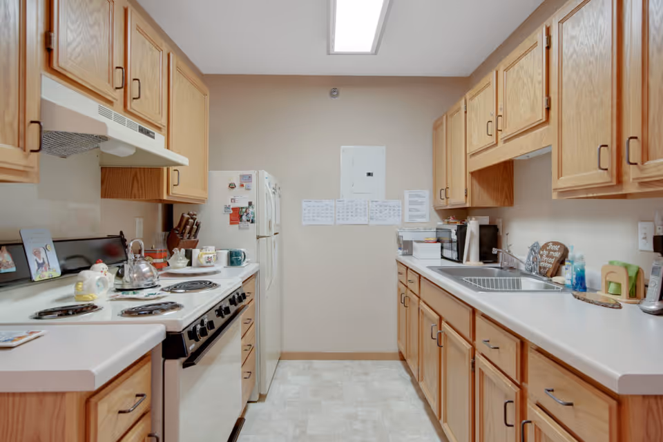 A well-lit kitchen with wooden cabinets on both sides, a white electric stove with a kettle and decorative items on the left, a white refrigerator at the end, and a sink with a drying rack and various kitchen items on the right countertop.