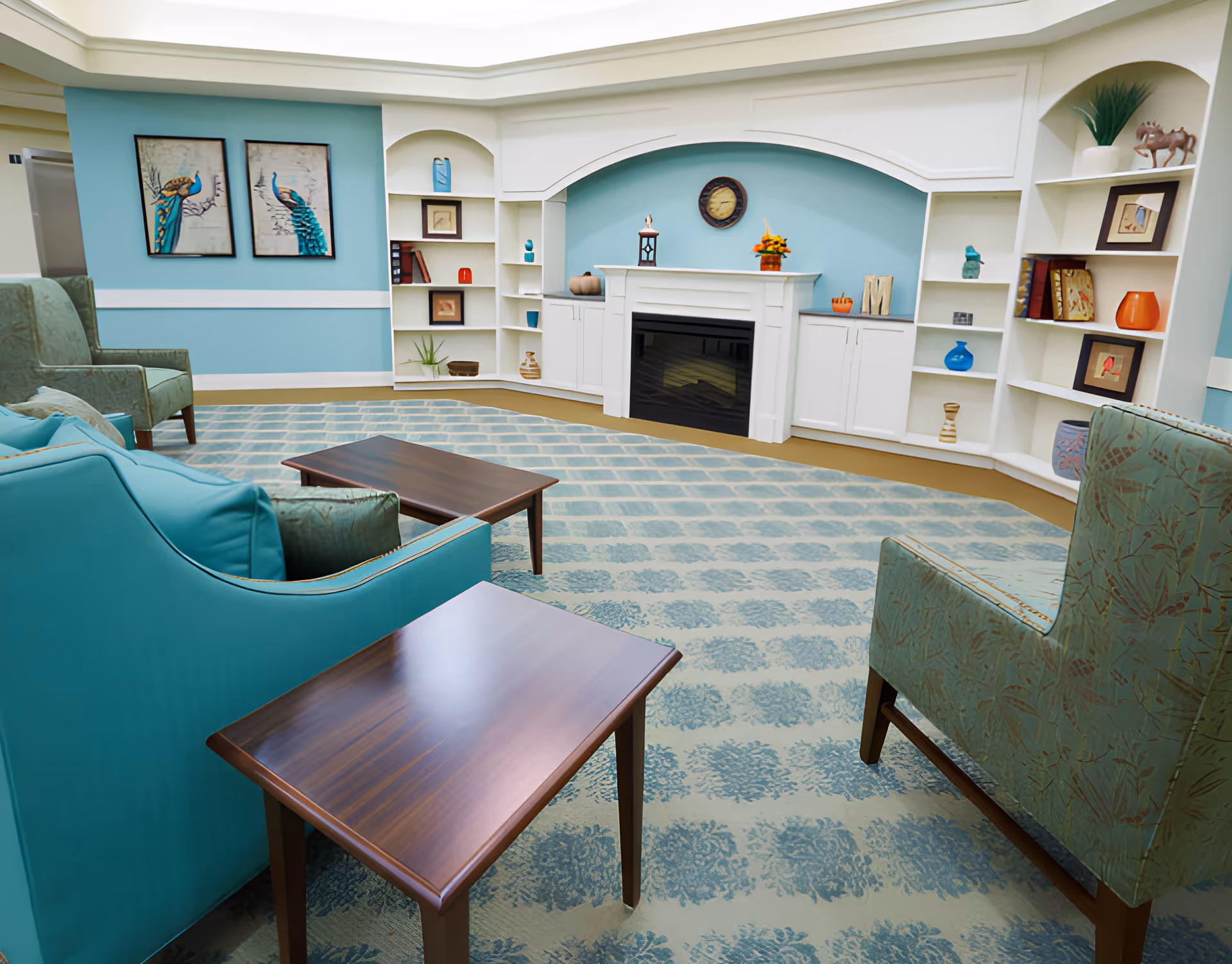 A cozy living room area with teal and green upholstered chairs and sofas, wooden coffee tables, a patterned carpet, and built-in white shelves surrounding a white fireplace. The walls are painted light blue with two framed peacock paintings on one side and various decorative items on the shelves.