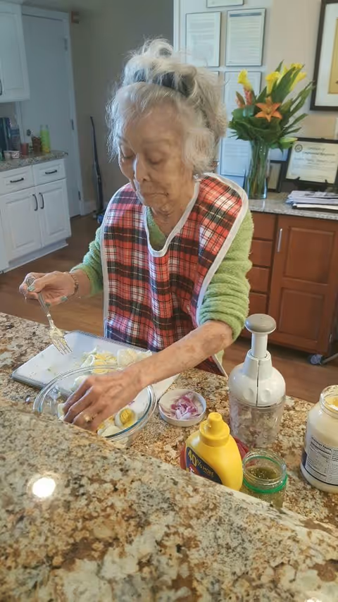An elderly woman wearing a red plaid bib is preparing food in a kitchen. She is placing sliced boiled eggs into a glass bowl on a granite countertop. Various kitchen items including mustard, mayonnaise, a small bowl with sliced onions, and a food chopper are visible on the counter. Behind her, there are wooden cabinets, a vase with flowers, and framed certificates on the wall.