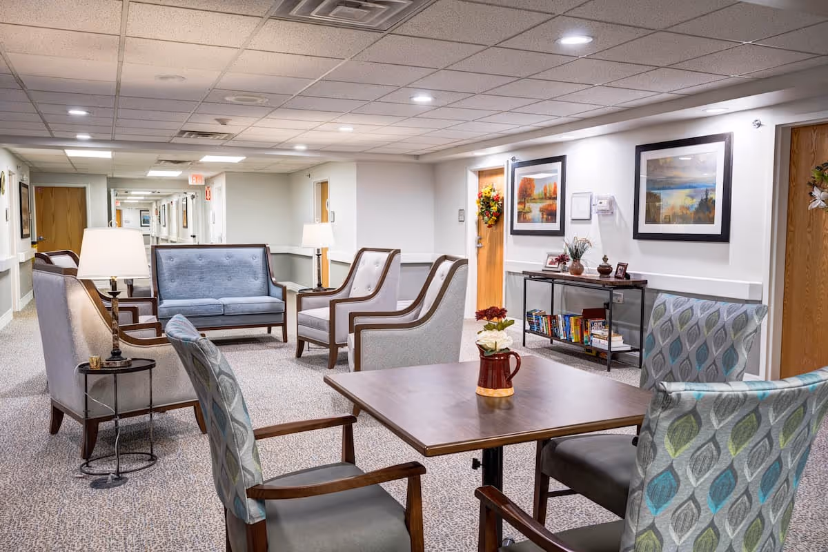 A well-lit common area in a senior living facility with comfortable seating including armchairs and a loveseat arranged around a wooden table. The walls are decorated with framed artwork, and a console table holds books and decorative items. The hallway with wooden doors extends into the background.