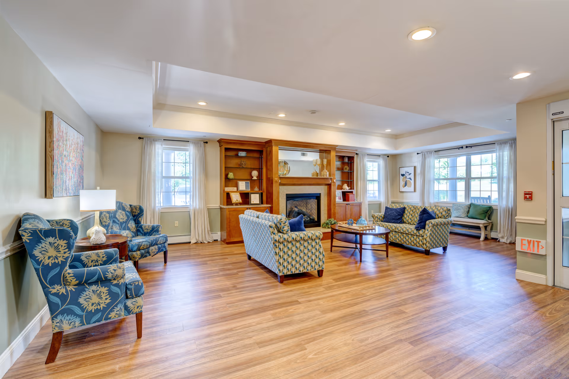 A spacious and well-lit living room with wooden flooring, featuring two patterned blue armchairs with a small side table and lamp between them on the left, two patterned sofas with blue cushions arranged around a round wooden coffee table in the center, and a wooden built-in shelving unit with a fireplace in the middle background. Large windows with sheer white curtains allow natural light to fill the room, and a bench with cushions is positioned near the windows on the right side.