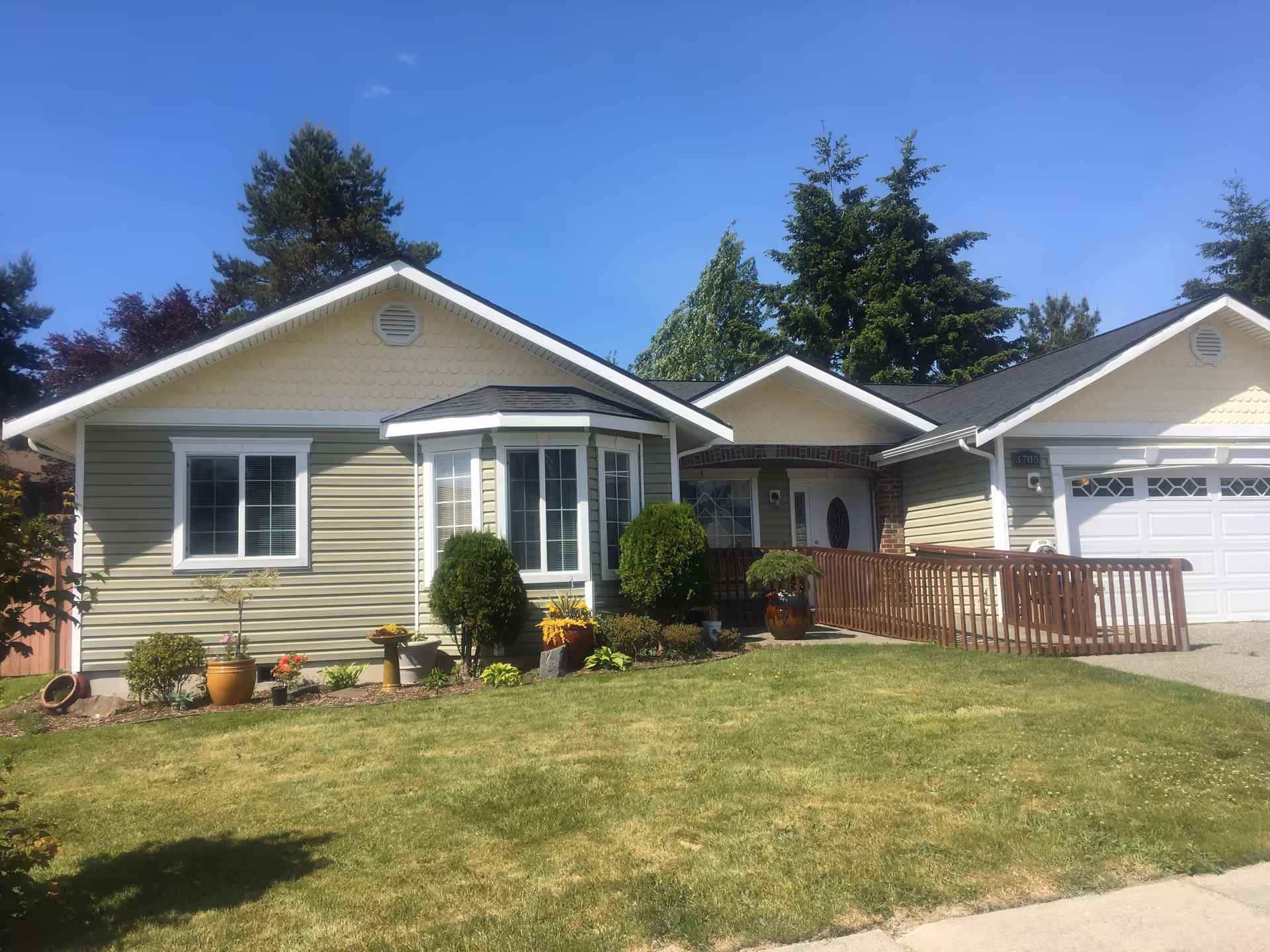 Single-story residential building with light yellow siding, white trim, and a dark roof. The front yard has a well-maintained lawn, various potted plants, shrubs, and a wooden ramp leading to the front door. Tall trees are visible in the background under a clear blue sky.