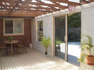 Sunlit outdoor patio area with a wooden deck, table, chairs, and sliding glass doors.