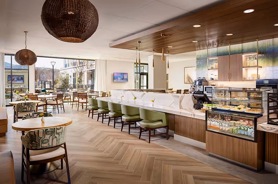 A bright and modern dining area with wooden floors and large windows letting in natural light. The space features a long marble counter with green cushioned chairs, round tables with patterned chairs, and a display case with food and beverages. Pendant lights hang from the ceiling, and there is a view of an outdoor patio area through the windows.
