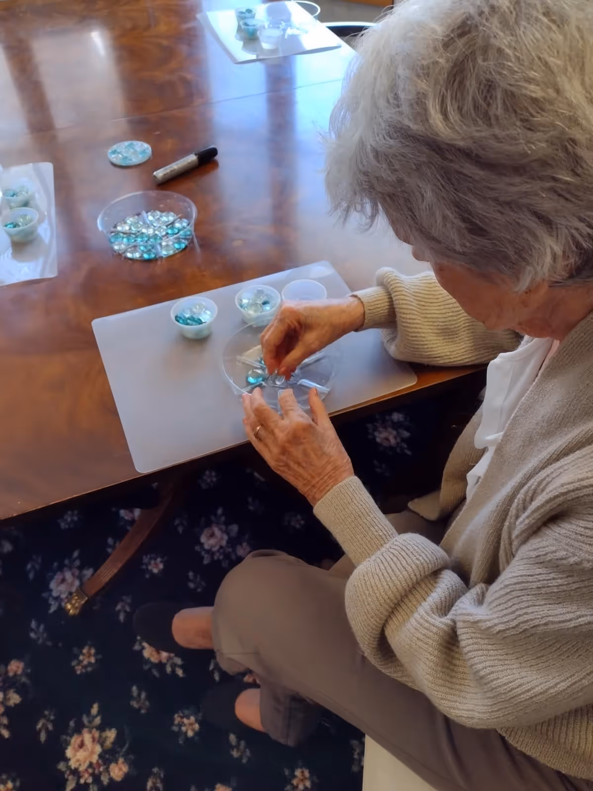 An elderly woman sitting at a wooden table engaged in a craft activity, sorting and arranging blue and clear decorative glass stones into small containers. She is wearing a beige cardigan and brown pants, and the table has a floral carpet underneath.