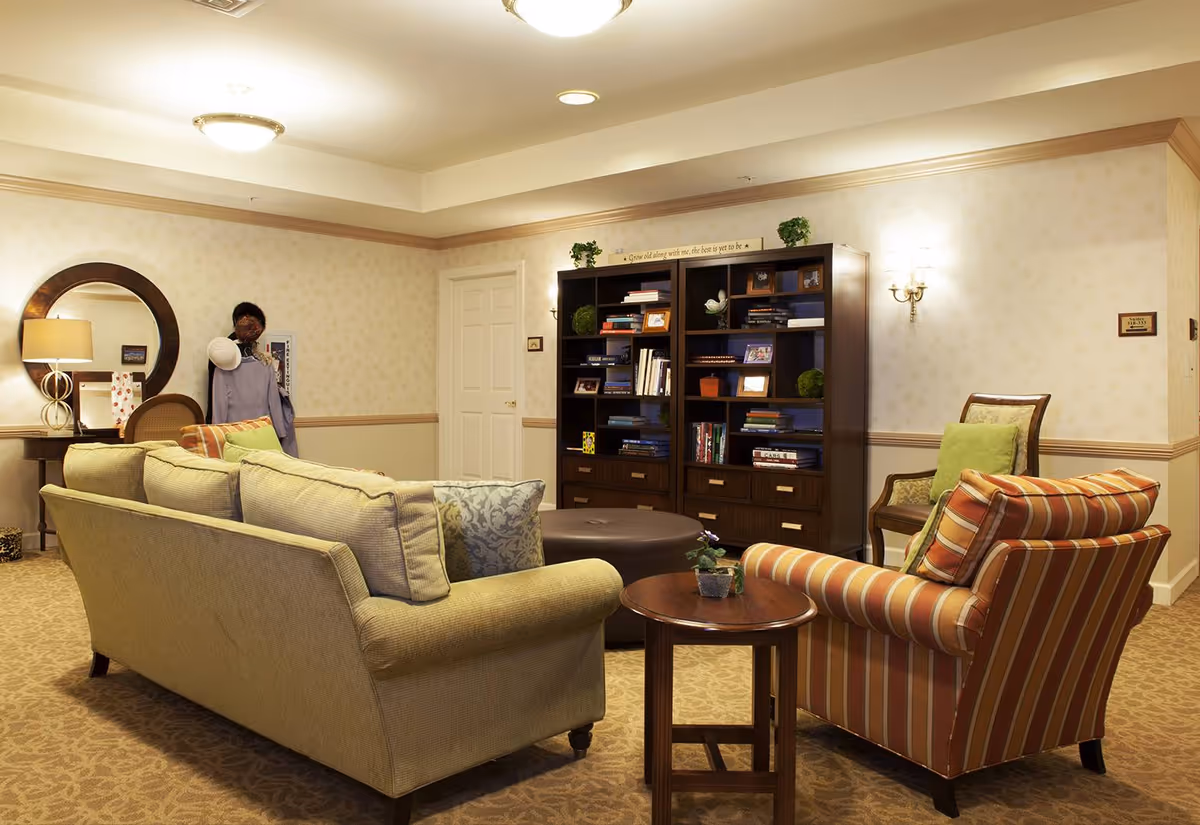 A cozy living room area with a beige sofa, a striped armchair, a round wooden side table with a small plant, and a large dark wood bookshelf filled with books and decorative items. The room has beige patterned carpet, wallpaper with a subtle pattern, a round mirror above a small table with a lamp, and soft lighting from ceiling and wall fixtures.