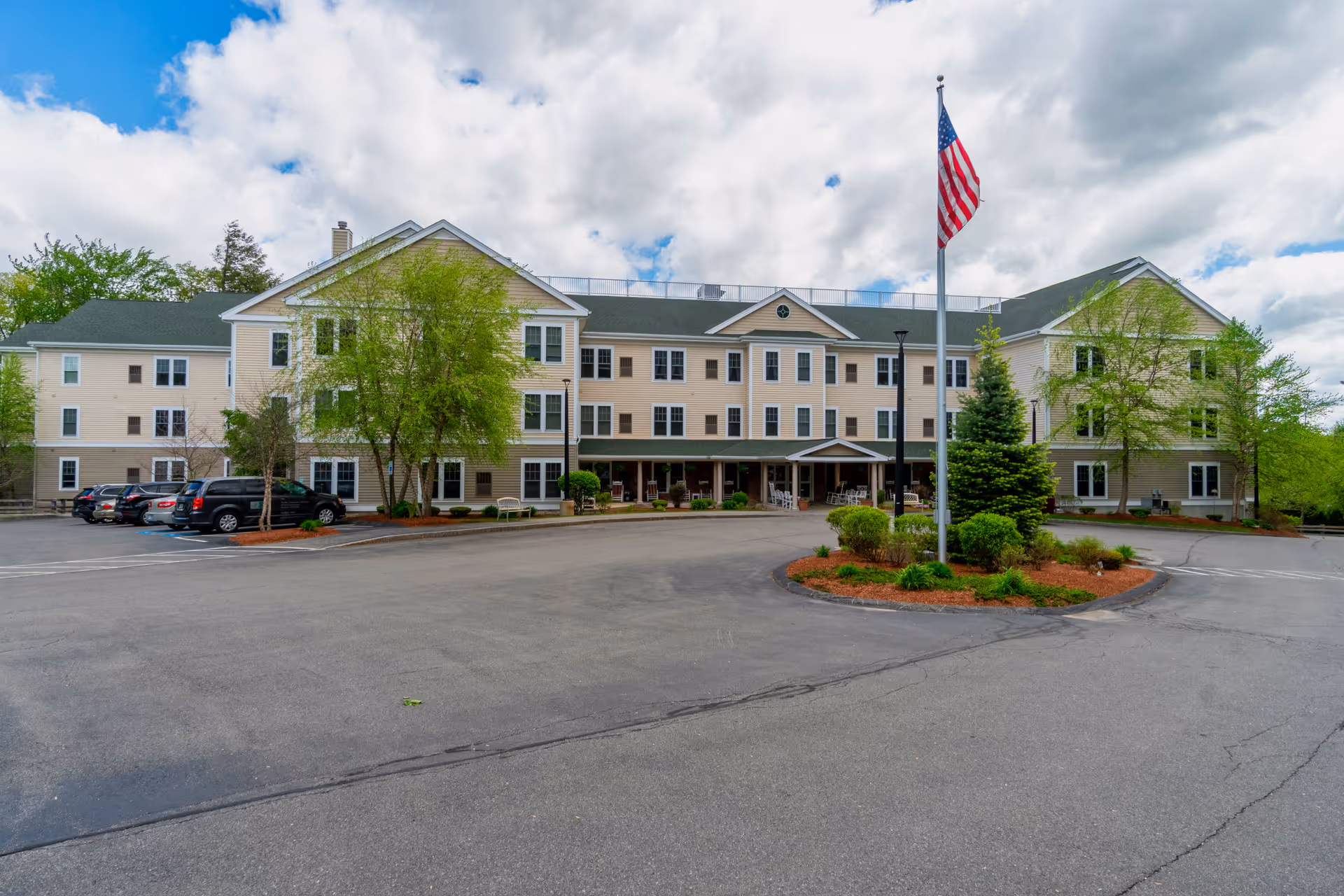 Exterior view of a three-story senior living facility building with beige siding and white trim. There is a circular driveway with a landscaped island in the center featuring an American flag on a flagpole and various shrubs and small trees. Several cars are parked along the left side of the building. The sky is partly cloudy.