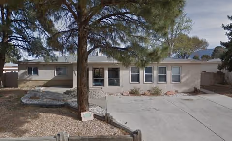 Single-story beige senior living facility with a concrete driveway and front entry, tree in foreground.
