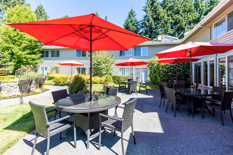 Outdoor patio area at a senior living facility with multiple round glass tables surrounded by wicker chairs. Each table has a large red umbrella providing shade. The patio is surrounded by greenery, including bushes, trees, and flowers, with a multi-story building in the background under a clear blue sky.
