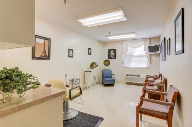 A bright room in a senior living facility with a hair salon chair and hair drying equipment on the left side, a blue armchair near the window at the back, and three wooden chairs with cushions along the right wall. The room has framed pictures on the walls, a window with white blinds and a valance, and a small wall-mounted TV in the corner.